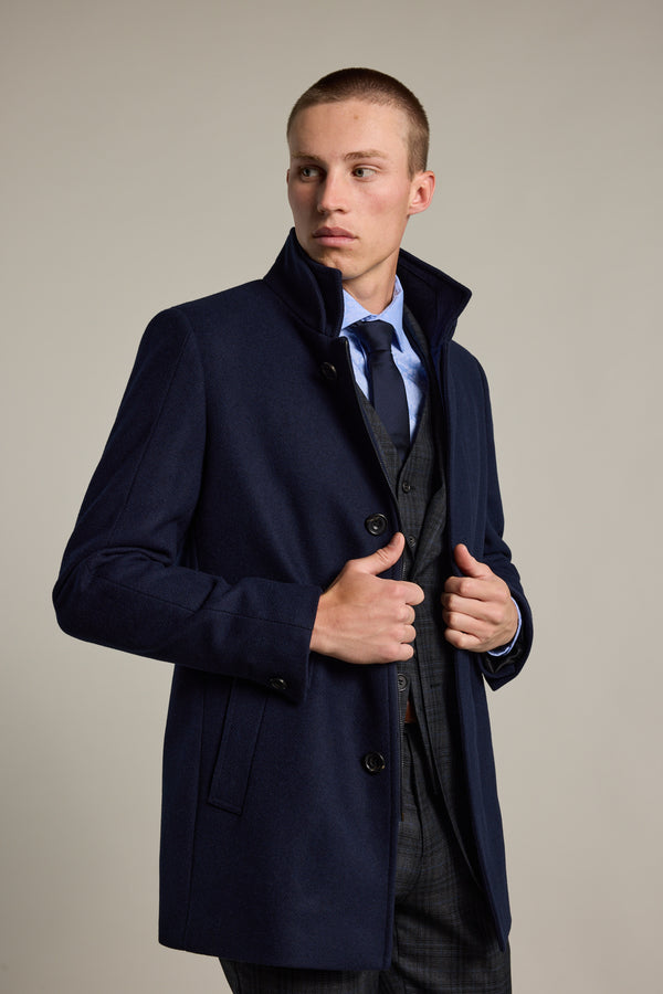 A young man in a navy blue Barkers Raymond Coat over a checked suit and blue shirt with tie stands facing left, adjusting his coat. He looks away from the camera against a plain, light background.