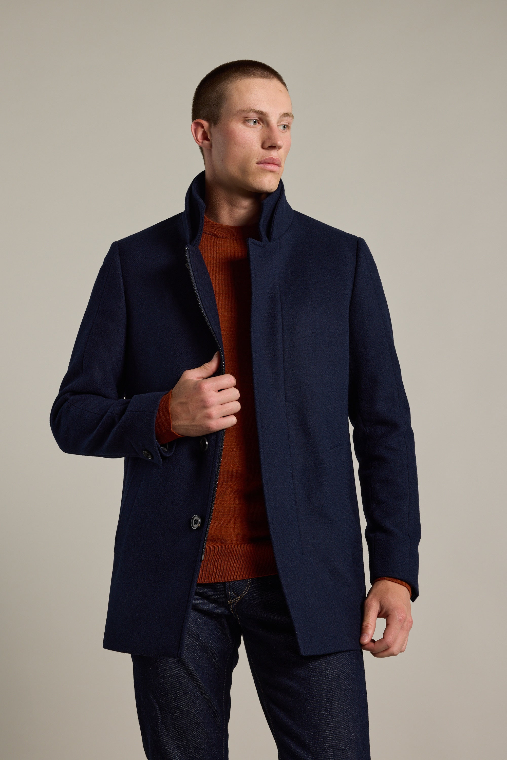 A man stands against a neutral background, wearing the Barkers Raymond Coat—a dark blue wool blend—over a rust sweater and dark jeans, gazing slightly to the side with one hand holding his coat.