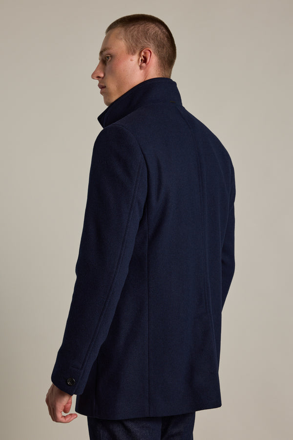 A man with short hair wears the Barkers Raymond Coat, a dark navy wool blend with turned-up collar, standing slightly turned and looking left against a plain light background.