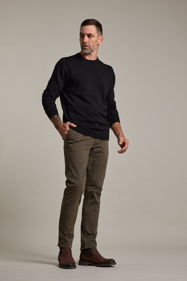 A man with short dark hair and a beard stands against a plain background, wearing a black long-sleeve sweater, Barkers Carnaby Chino in olive green, and brown boots. He looks slightly to the side with a neutral expression.