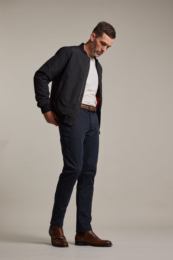 A man stands against a plain light background, wearing Barkers Carnaby Chino pants styled with a black bomber jacket, light gray shirt, brown belt, and brown Chelsea boots. He looks down with his hands in his pockets.