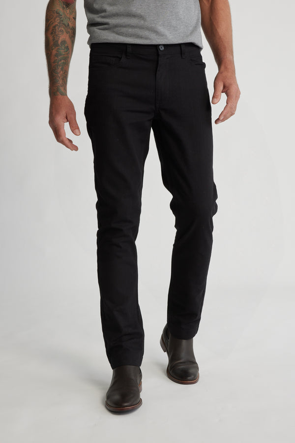 A man in a gray T-shirt, Barkers Oxmor 5 Pkt Pant O in black (taper fit), and dark brown boots stands against a white background. His arms are relaxed at his sides, with visible tattoos on one arm.