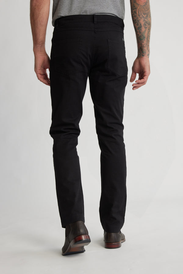 A person stands facing away in a grey t-shirt, Barkers Oxmor 5 Pkt Pant O (taper fit), and brown dress shoes, revealing a tattooed left arm against a neutral background.