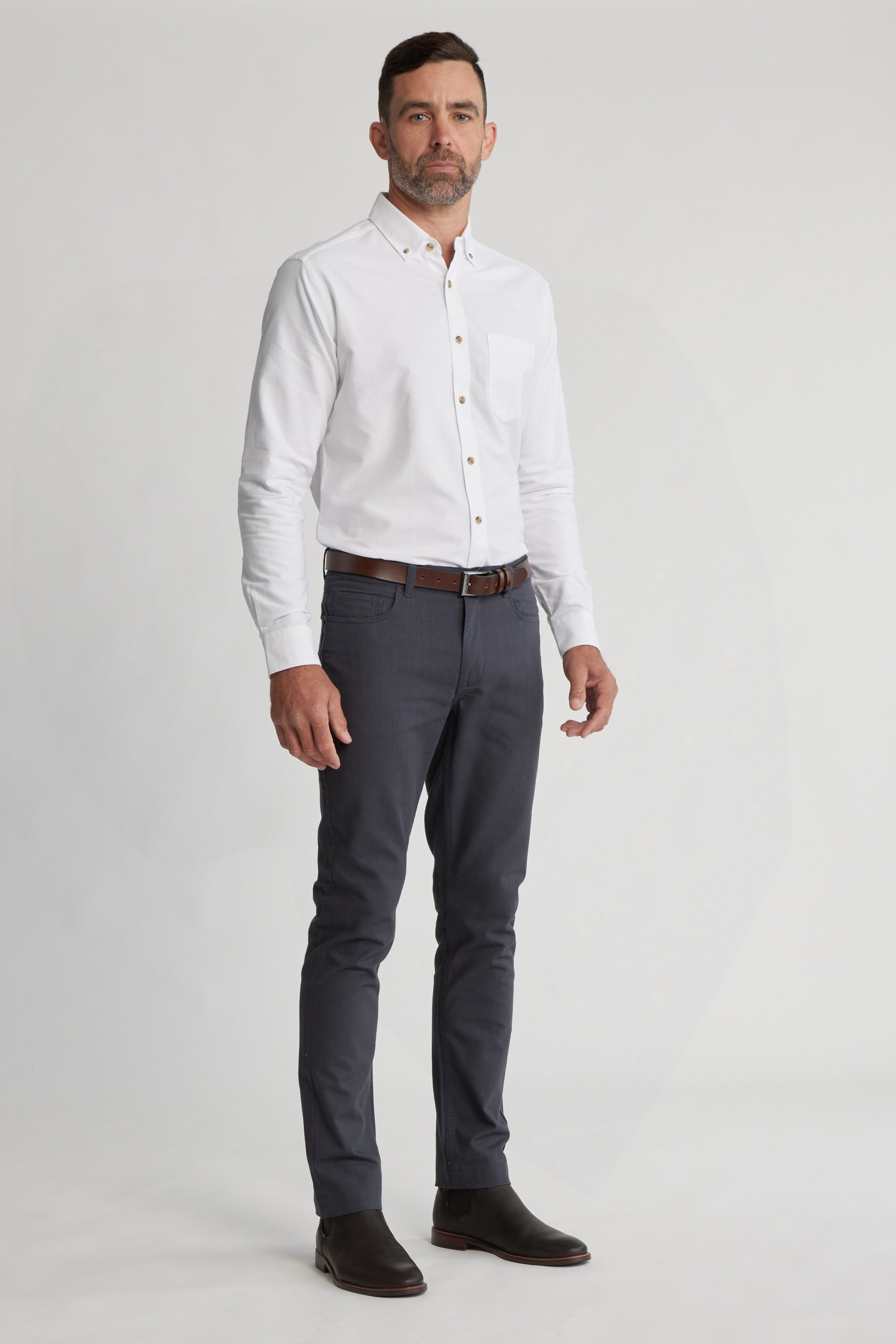 A man with short hair and a trimmed beard stands against a plain light background, wearing Barkers Oxmor 5 Pkt Pant O in dark gray with a white shirt, brown belt, brown shoes, and a neutral expression.