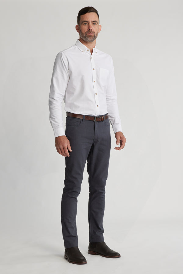A man with short hair and a trimmed beard stands against a plain light background, wearing Barkers Oxmor 5 Pkt Pant O in dark gray with a white shirt, brown belt, brown shoes, and a neutral expression.