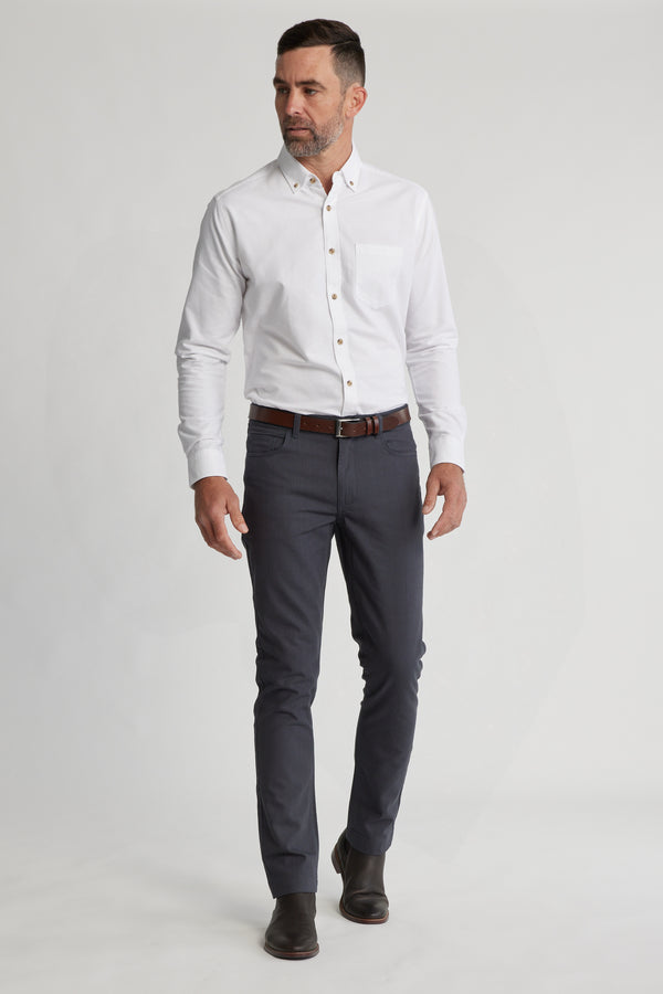 A man in a white button-up shirt and Barkers Oxmor 5 Pkt Pant O stands against a plain light background, paired with a brown belt and shoes, looking slightly to the side with a neutral expression.