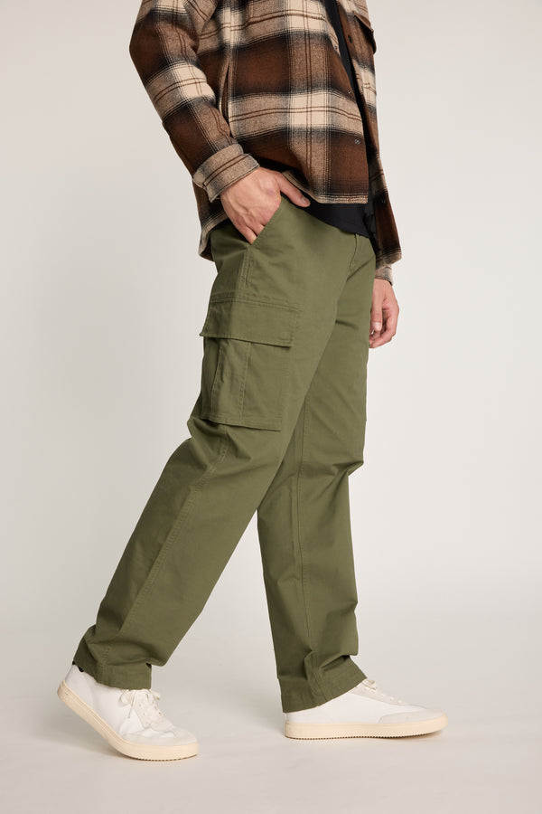 Someone stands against a neutral background wearing Barkers Barnard Cargo Pant in olive green twill cotton, paired with a black shirt, brown plaid jacket, and white sneakers.