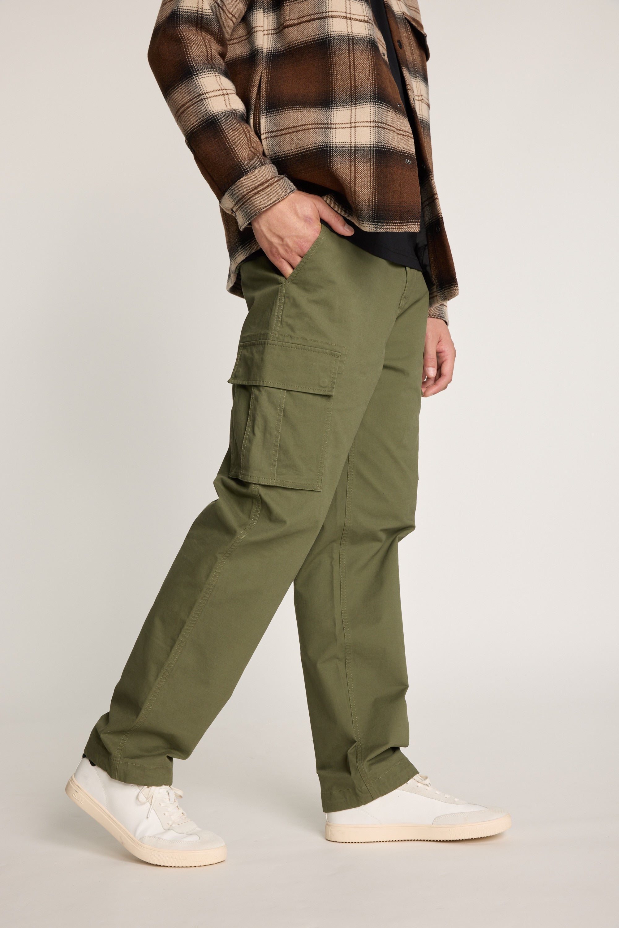 Someone stands against a neutral background wearing Barkers Barnard Cargo Pant in olive green twill cotton, paired with a black shirt, brown plaid jacket, and white sneakers.