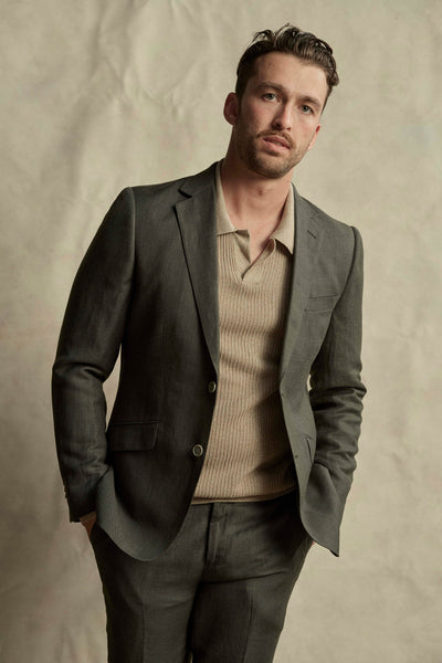 A man with short dark hair and a trimmed beard wears the Barkers Inverness Suit Jacket over a beige knit polo, standing with hands in his pockets against a neutral, textured backdrop and looking slightly to the side.