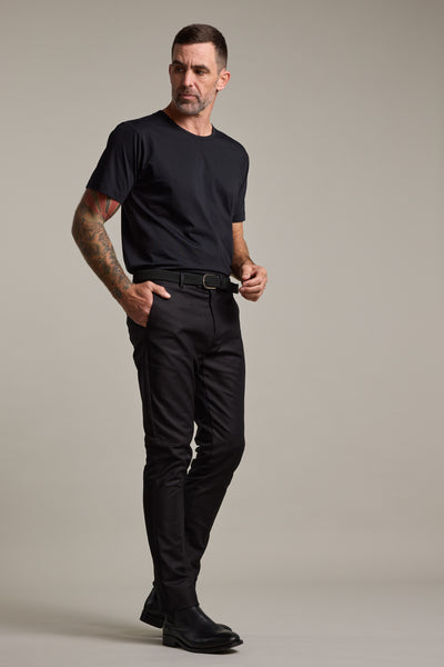A man with short hair and arm tattoos stands against a plain backdrop, wearing Barkers Lyndon Cotton Pants, a black t-shirt, belt, and black shoes in a smart casual style, looking slightly to the side with one hand on his belt.