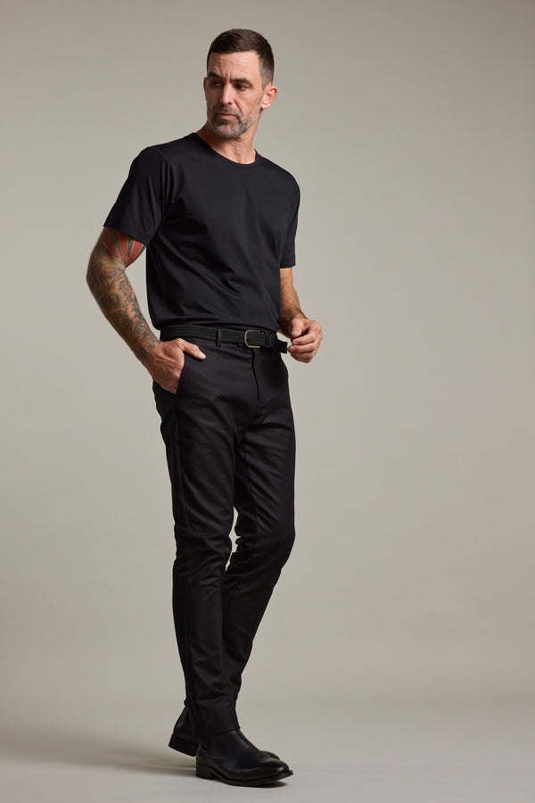 A man with short hair and arm tattoos stands against a plain backdrop, wearing Barkers Lyndon Cotton Pants, a black t-shirt, belt, and black shoes in a smart casual style, looking slightly to the side with one hand on his belt.