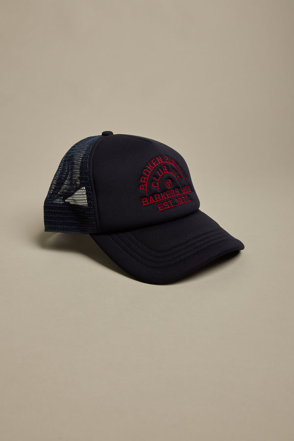 The Barkers Piston Club Trucker Cap, featuring a black mesh design with a red embroidered logo on the front, is displayed against a neutral beige background.