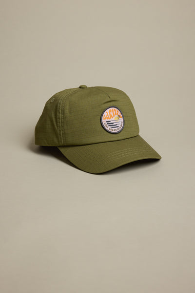 The Barkers Longboard Cap by Barkers is an olive green snapback featuring a round, colorful embroidered "CACKLE" patch with a sunset over mountains on the front. It is displayed on a light beige background.