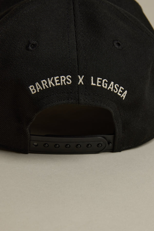 Close-up of the back of the black Legasea Logo Snapback Cap with an adjustable strap. White embroidered Legasea and BARKERS X LEGASEA logos appear above the strap for a customizable fit.