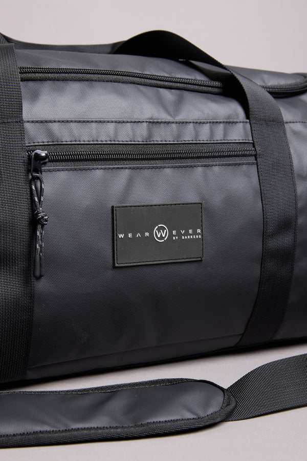 A close-up of the WearEver Transit Holdall, a black, water-resistant duffel bag with a zipper pocket, shoulder strap, visible stitching, WEAR EVER BY LAMAR label in white, and a convenient sneaker pocket.