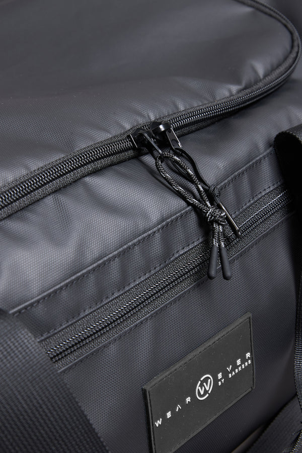 Close-up of the WearEver Transit Holdall, a black duffel bag with visible zipper pulls and a WearEver label. Made from textured, water-resistant fabric, it offers detailed stitching and versatile storage for travel essentials.
