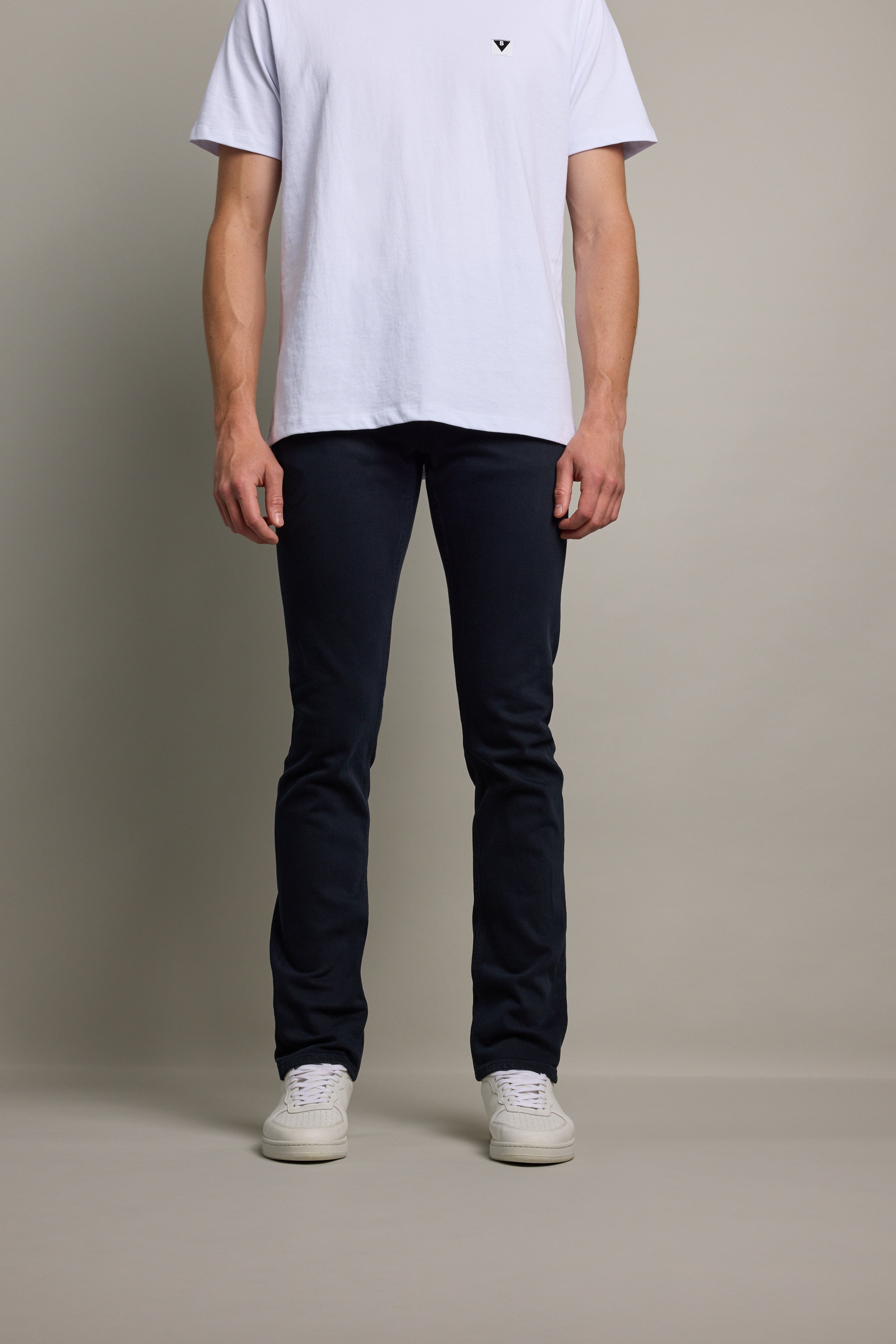 A person stands against a plain background wearing a white t-shirt, Barkers Rambler Slim Coloured Jean made from regenerative cotton by Saitex Vietnam, and white sneakers. The image shows the person from shoulders to feet.