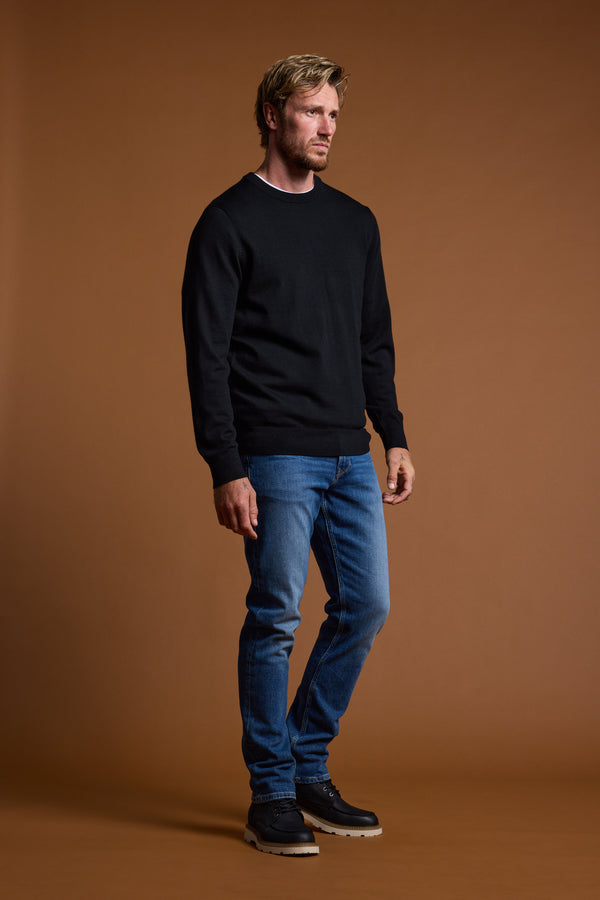 Man in a Barkers Skyland ZQrx Merino Crew, black sweater and blue jeans, stands by brown backdrop.