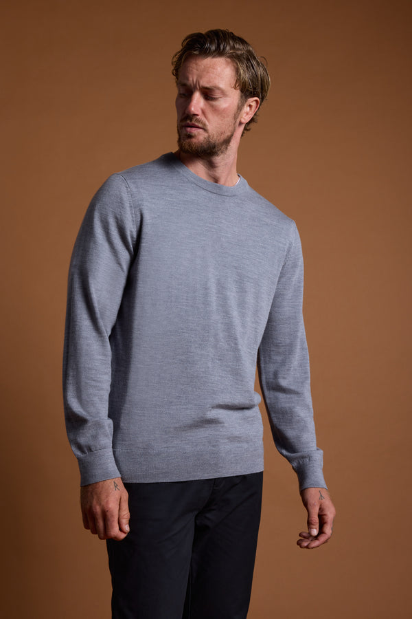 A man in a light gray Barkers Skyland ZQrx Merino Crew and black pants stands before a plain brown background, gazing slightly left with a neutral expression.