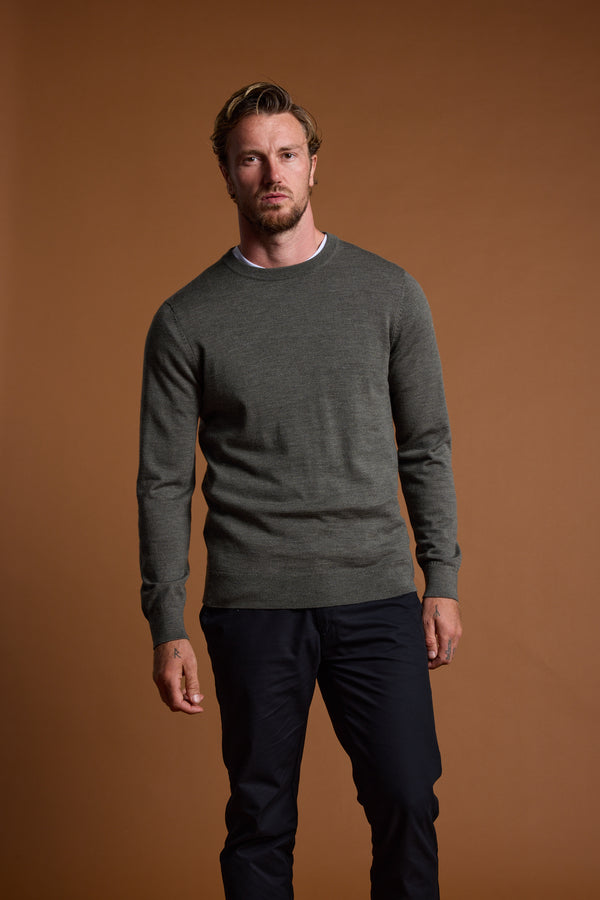 A man with short, wavy hair and a trimmed beard stands against a plain brown background, wearing the Barkers Skyland ZQrx Merino Crew sweater over a white T-shirt and dark pants.