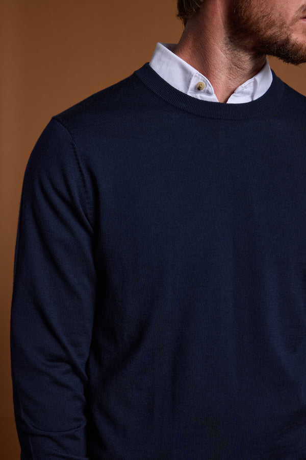 A man wears a Barkers Skyland ZQrx Merino Crew sweater in navy, made from sustainable New Zealand merino wool, layered over a white collared shirt. Set against a plain brown background, only his torso and part of his face and neck are visible.