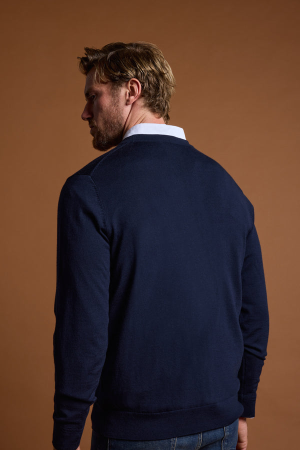 A man with light brown hair and a beard stands with his back to the camera, wearing a navy Skyland ZQrx Merino Crew sweater by Barkers over a white collared shirt. The background is plain brown.