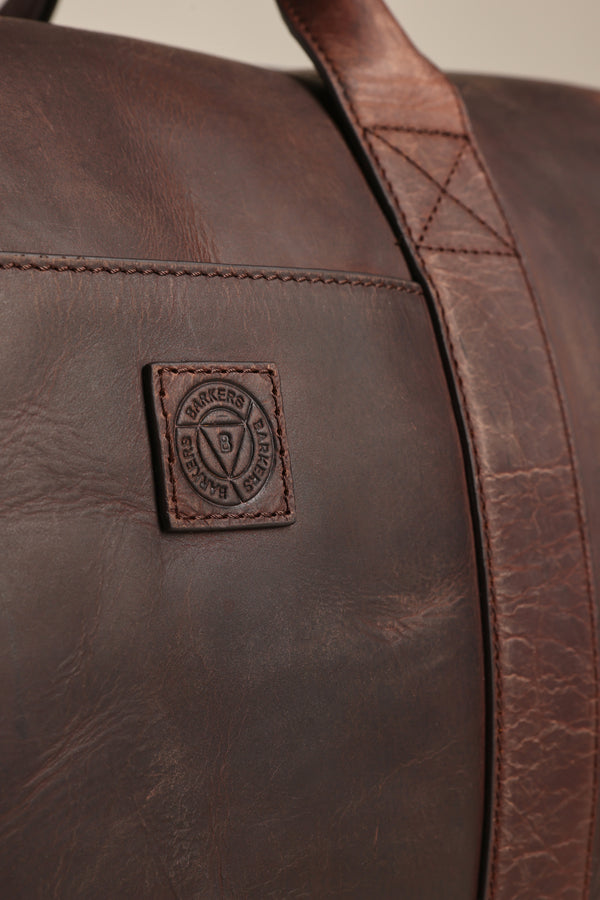 Close-up of the Costello Leather Weekender by Barkers, a brown leather bag with a stitched square patch displaying a circular embossed logo and text, plus a sturdy, visibly stitched leather strap.