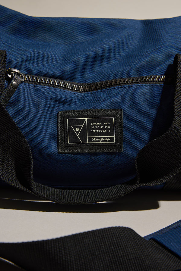 A close-up of the Barkers Canvas Duffle Bag in blue cotton canvas with a black zipper and strap. A stitched black label with white text and geometric logo is seen on the durable outer pocket of this Barkers bag.