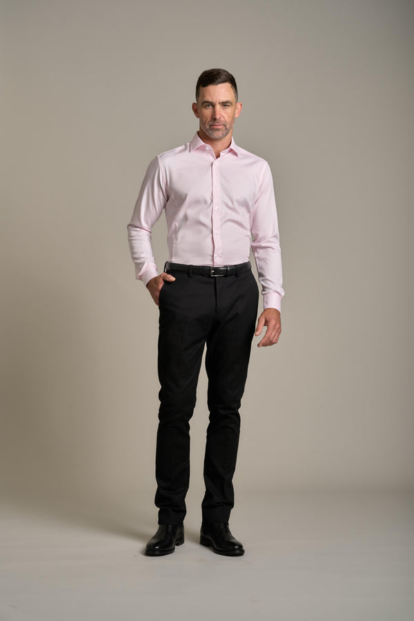 A man stands against a plain light background wearing a Barkers Tribeca Reg Cuff Shirt in light pink, black pants, and black shoes, with one hand in his pocket and a neutral expression.
