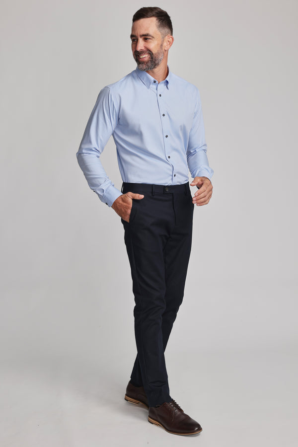 A man with short dark hair and a beard smiles as he walks in a Barkers Verona Shirt—tailored fit, long sleeve, 100% cotton—paired with black trousers and brown dress shoes against a plain light background.
