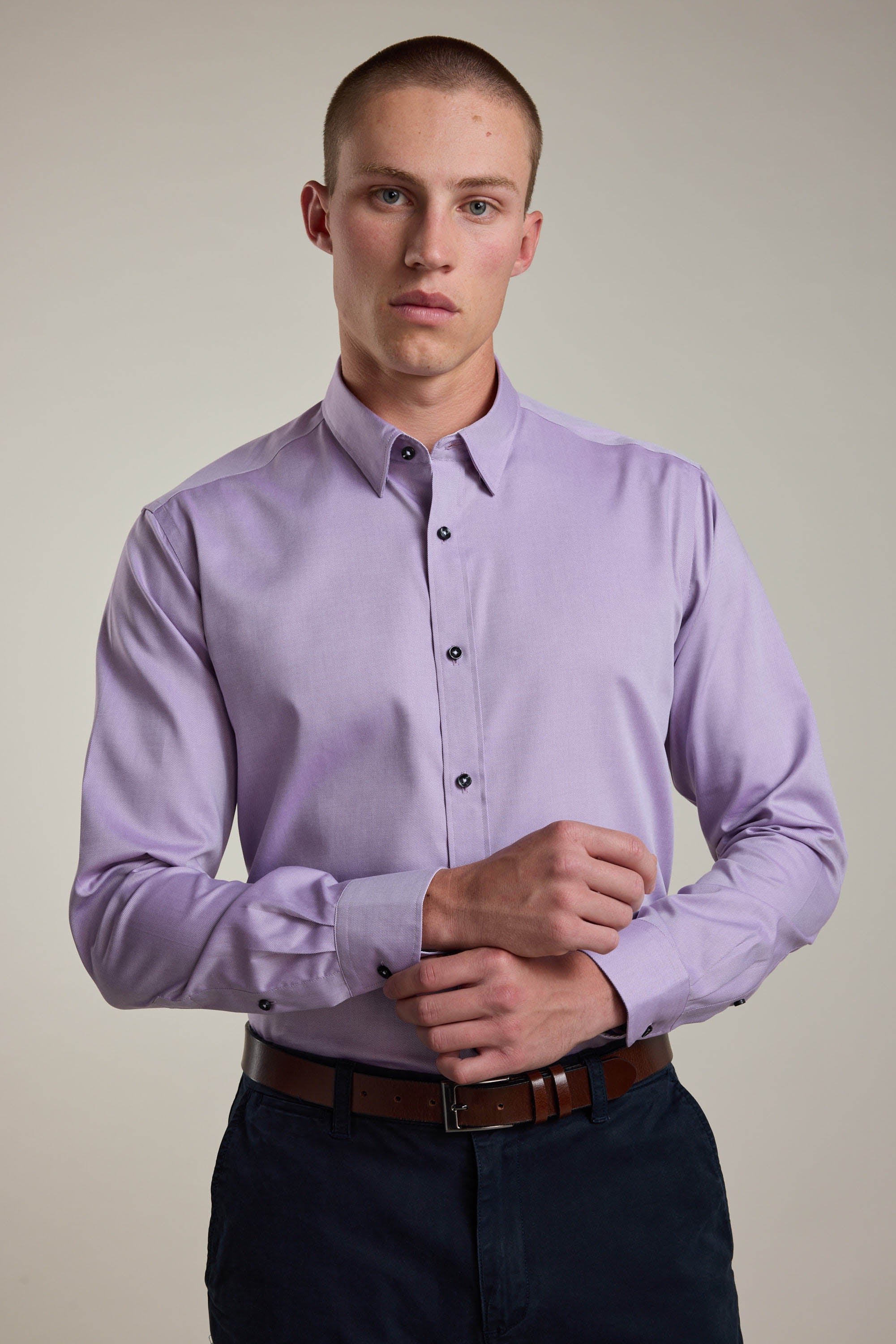 A young man with short hair wears the Barkers Verona Shirt, a tailored fit, long sleeve business shirt in light purple 100% cotton. Paired with dark pants and a brown belt, he stands against a plain background, adjusting his cuff and looking at the camera.