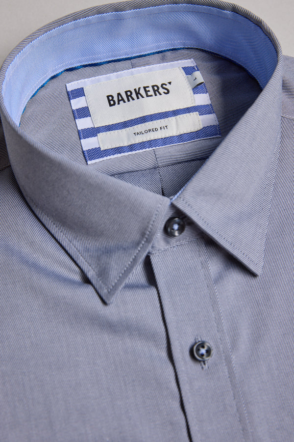The Barkers Verona Shirt is a neatly folded light grey long sleeve business shirt with a pointed collar, blue buttons, and an inner label reading "Barkers Tailored Fit" on a white and blue striped background.