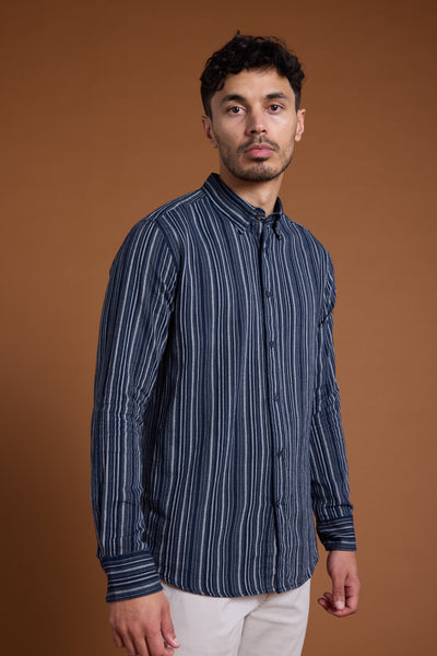 A man with short dark hair and a trimmed beard wears the Barkers Scott Stripe Shirt—a navy and white tailored fit shirt made from responsibly sourced cotton—paired with light pants, standing against a plain brown background.