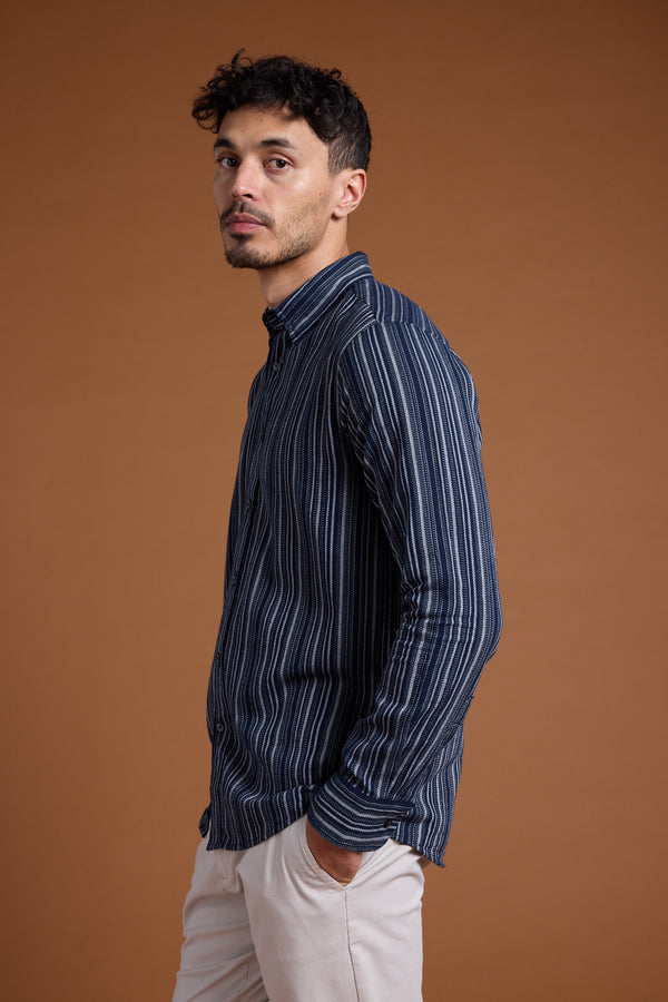 A man with short curly dark hair wears the Barkers Scott Stripe Shirt, made from responsibly sourced cotton, and light beige pants. He stands against a plain brown background with one hand in his pocket, gazing to the side.