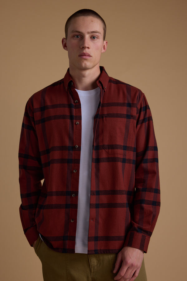 A man wears the Barkers Durness Check Shirt, made from responsibly sourced cotton in a rust check pattern, over a white tee and olive green pants, standing against a beige background with a neutral look.