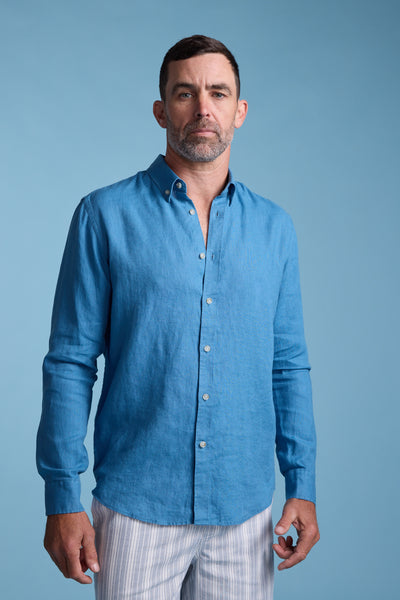 A man with short dark hair and a beard wears the Barkers Dorset Linen Shirt in blue with light-colored striped pants, standing against a plain light blue background.