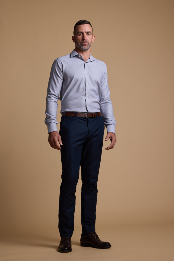 A man with short hair and a beard stands against a plain beige background wearing the Barkers Eaton Jacquard Business Shirt, tailored fit in light blue, paired with dark blue trousers, a brown belt, and brown dress shoes.
