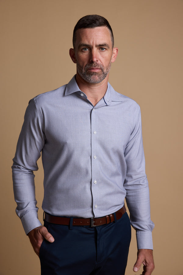 A man with short hair and a trimmed beard wears a light blue Barkers Eaton Jacquard Business Shirt and dark blue pants, standing against a plain beige background with a serious expression.
