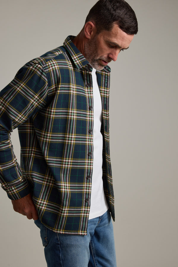 A man models the Barkers Brennan Check Overshirt in green and yellow plaid, made from responsibly sourced cotton, worn over a white T-shirt and blue jeans, standing with one hand in his pocket against a neutral background.