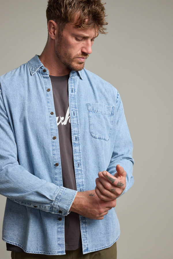 A man wears the Barkers Ruxton Denim Shirt in a classic fit, styled open over a dark tee, looking down while holding his right wrist with his left hand against a plain background.