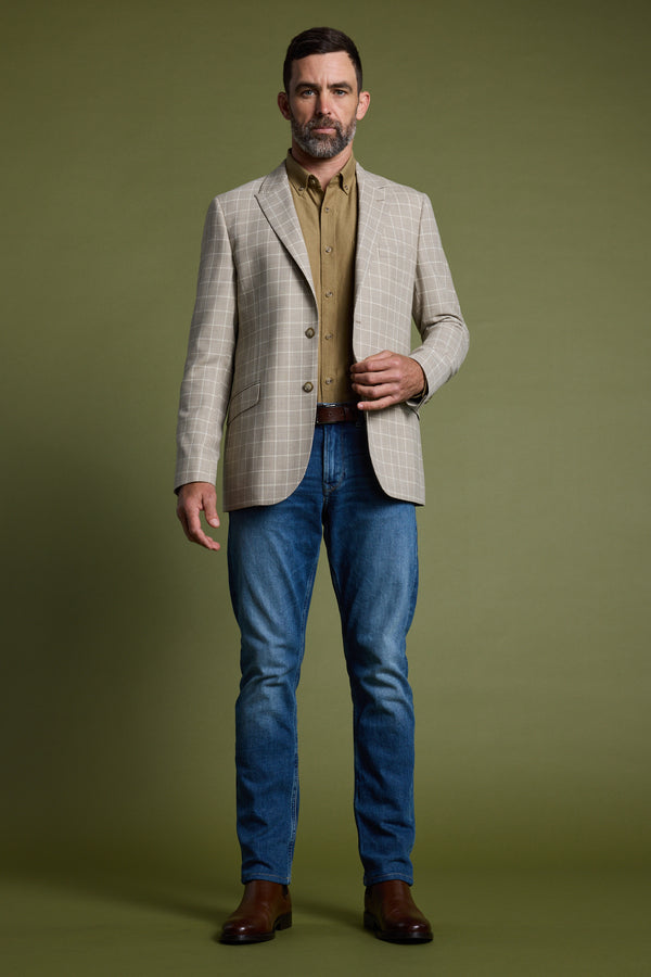 A man with short brown hair and a beard stands against an olive green background, wearing the Barkers Prestwick Check Blazer, a light brown shirt, blue jeans, and brown boots.