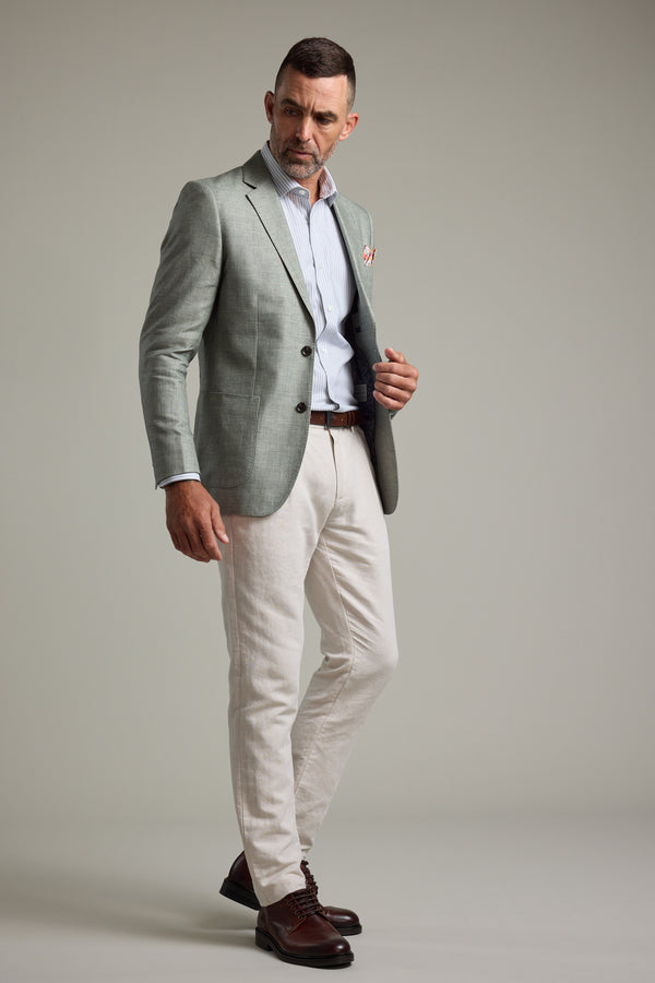 A man stands confidently against a plain light gray background wearing the Barkers Marion Texture Blazer, a striped dress shirt, beige trousers, brown belt, and brown dress shoes—a perfect summer occasion outfit.