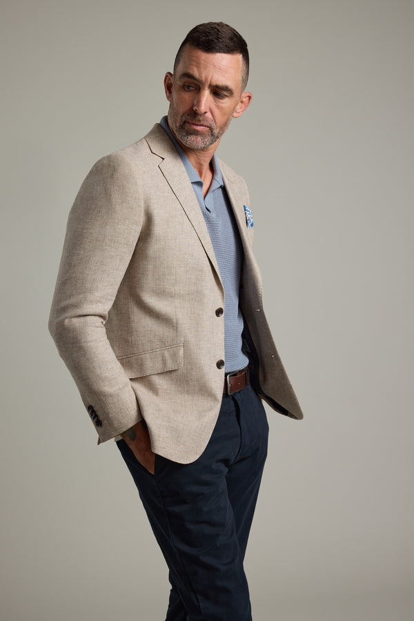 A man with short dark hair and a beard wears the Welland Texture Blazer by Barkers over a light blue shirt, dark pants, and a brown belt, standing with one hand in his pocket and looking to his left against a plain background.