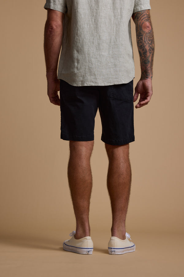 A man with a tattooed right arm faces away from the camera, wearing Barkers Tidal GD Utility Short in black, a beige garment-dyed cotton short-sleeve shirt, and white slip-on shoes against a plain beige background.