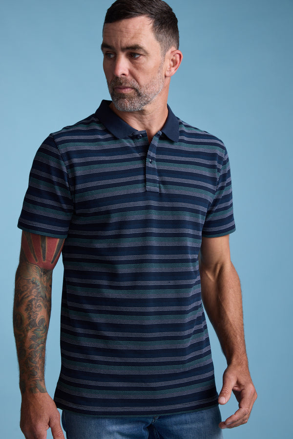A man with short dark hair and a beard wears the Barkers Dalton Stripe Polo with jeans, standing against a light blue background. His tattooed arm rests at his side as he looks right—a stylish pick for your summer wardrobe.