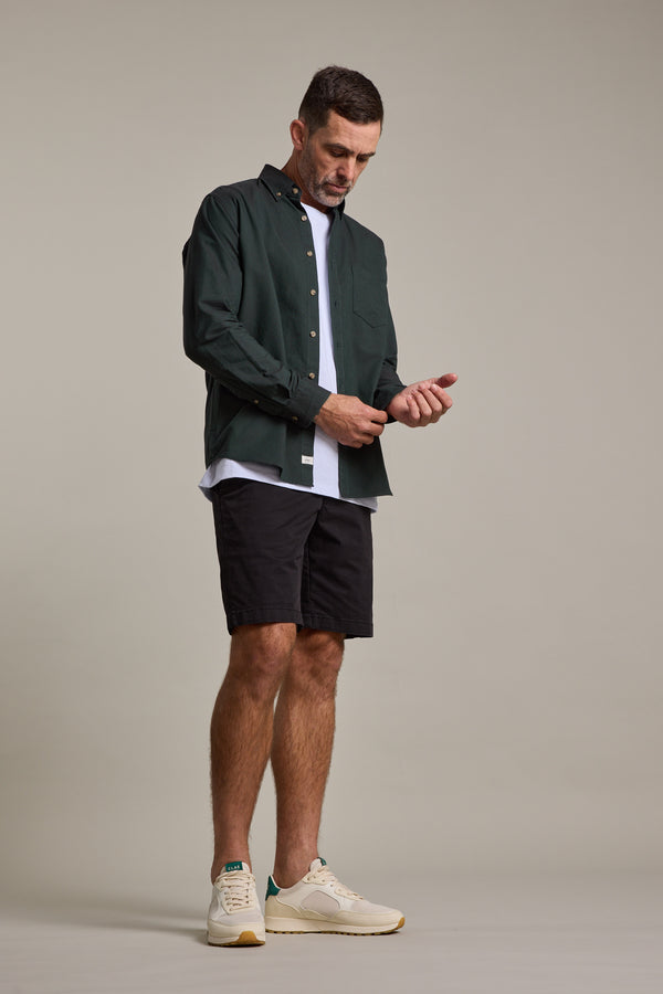 A man adjusts the cuff of his dark green button-up shirt worn over a white tee, paired with Barkers Kingly Chino Shorts in black and cream sneakers, set against a plain light background.