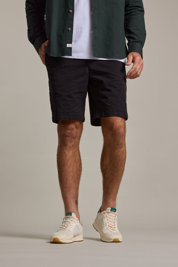 A man, cropped from the shoulders down, stands against a plain background wearing a dark green shirt over a white tee, Barkers Kingly Chino Short, and beige sneakers with white soles.