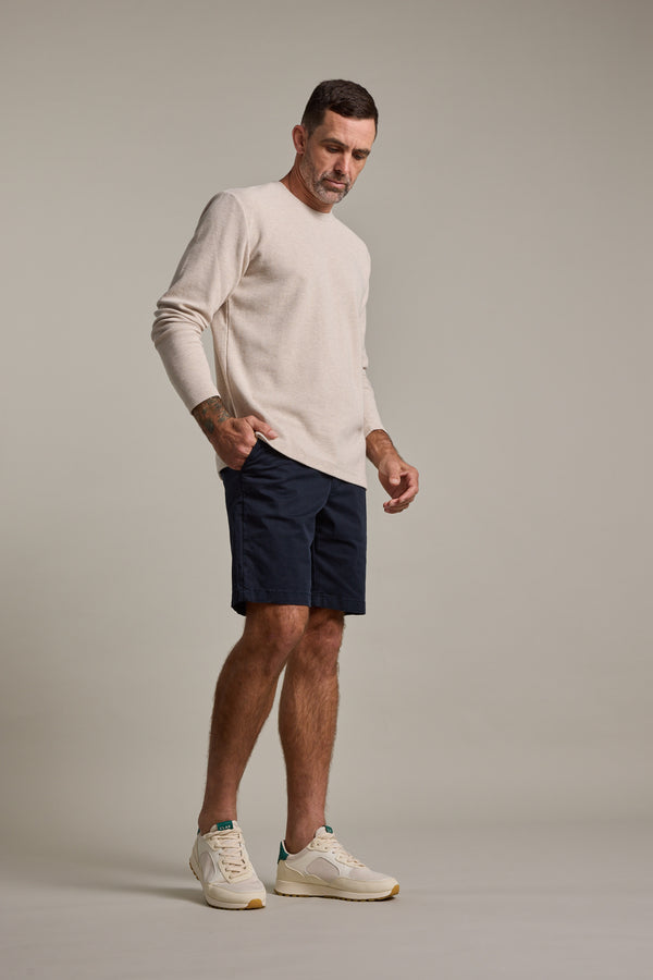 A man with short hair and a forearm tattoo stands against a plain background, wearing a light beige long-sleeve shirt, Barkers Kingly Chino Short in dark navy, and white sneakers, looking down with a relaxed expression.