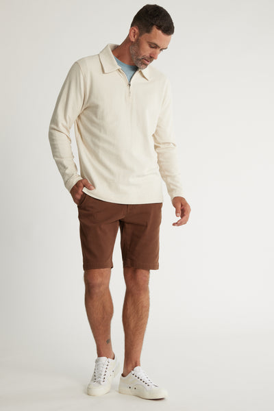 A man stands against a plain background wearing Barkers Thomas GD Walk Shorts, a cream zip-collar pullover over a light blue shirt, and white sneakers, looking down with one hand in his pocket.