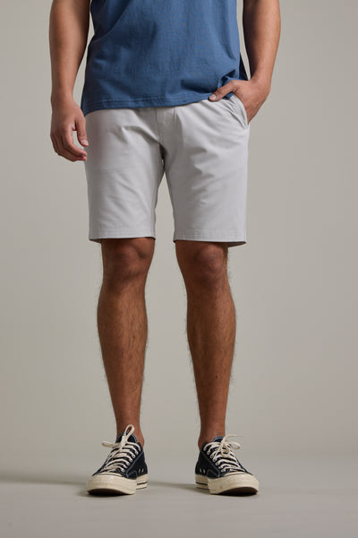 A person stands with one hand in their pocket, wearing a blue T-shirt and Barkers Hutchinson Walkshort light gray quick-dry shorts, paired with black sneakers featuring white laces. Image cropped from shoulders to feet.